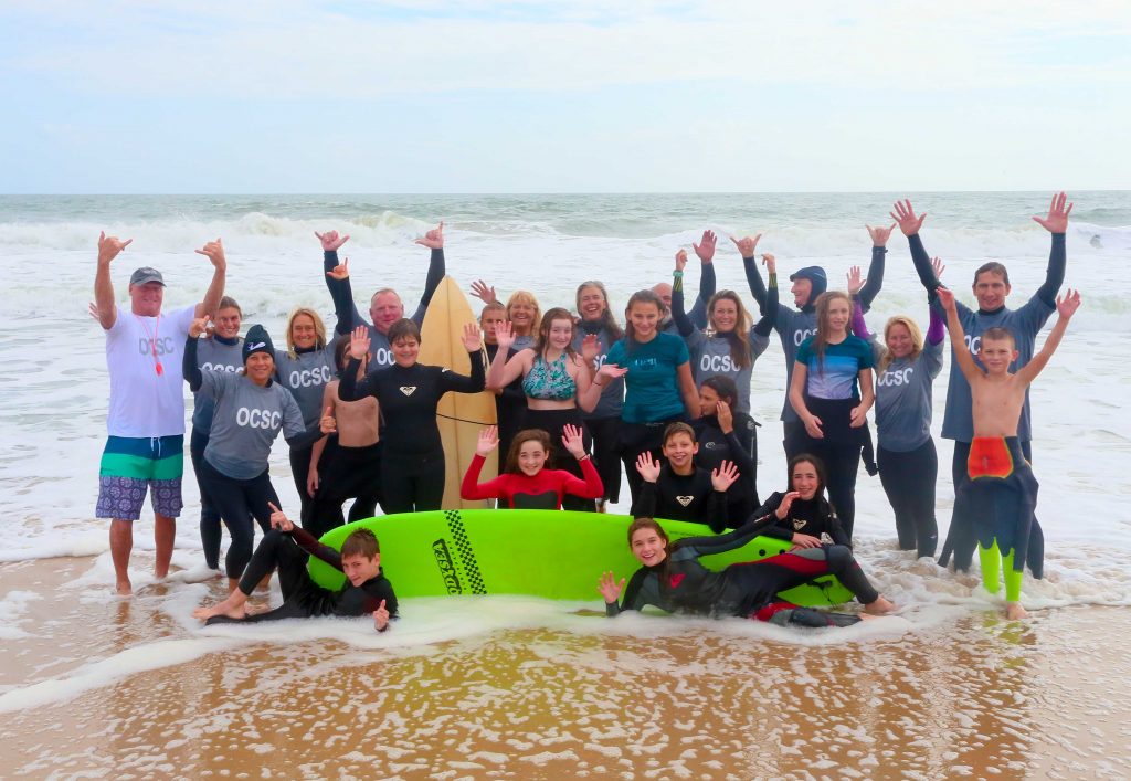 group of mostly kids in wetsuits in front of surboard on surf beach