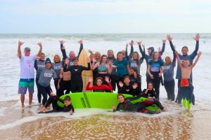 group of mostly kids in wetsuits in front of surboard on surf beach